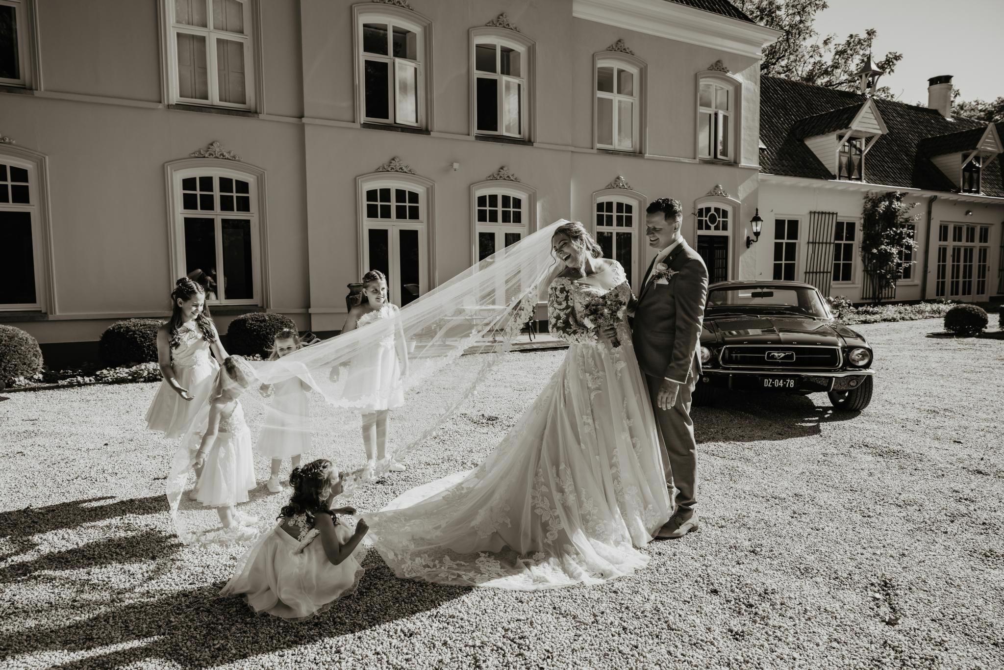 Real Bride Eline – bruid en bruidegom lachen samen terwijl bruidsmeisjes speels de lange sluier vasthouden; Eline draagt een romantische A-lijn trouwjurk met verfijnd kant en volumineuze rok, vastgelegd bij een statige locatie met klassieke auto voor een tijdloos en liefdevol moment.